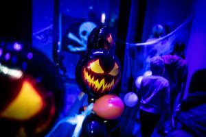 Halloween-teemainen kuva, jossa ilmapalloja ja kurpitsalyhdyn muotoisia ilmapalloja violetinsinisessä kuvassa. Taustalla nuoria, jotka ovat sumeana. Näkyy myös pääkallolippu ja hämähäkinseittiä. Koristeltu Willi Länsi -huvikeskuksen tila Himoksella.