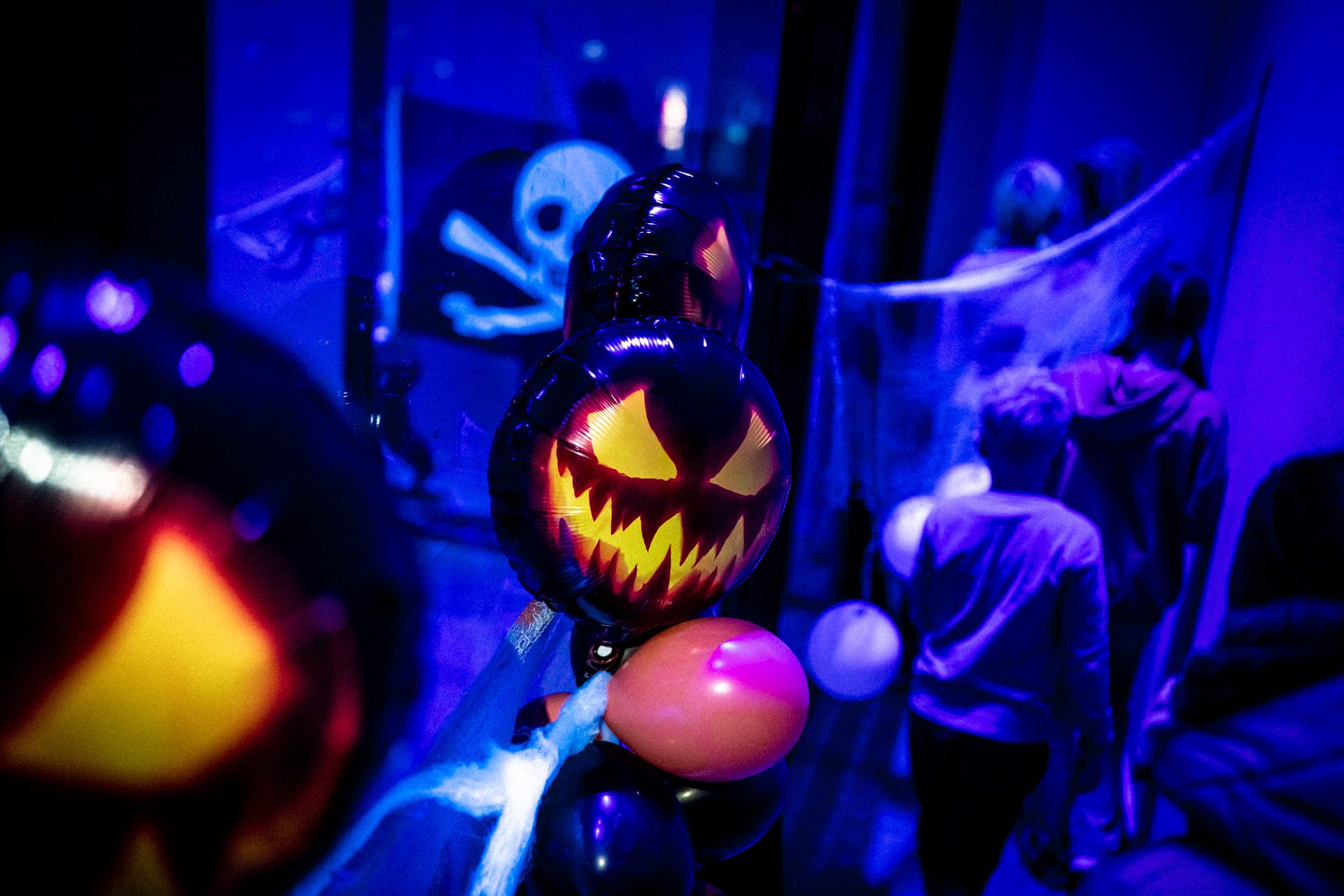 Halloween-teemainen kuva, jossa ilmapalloja ja kurpitsalyhdyn muotoisia ilmapalloja violetinsinisessä kuvassa. Taustalla nuoria, jotka ovat sumeana. Näkyy myös pääkallolippu ja hämähäkinseittiä. Koristeltu Willi Länsi -huvikeskuksen tila Himoksella.