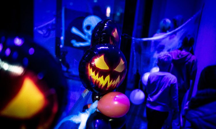 Halloween-teemainen kuva, jossa ilmapalloja ja kurpitsalyhdyn muotoisia ilmapalloja violetinsinisessä kuvassa. Taustalla nuoria, jotka ovat sumeana. Näkyy myös pääkallolippu ja hämähäkinseittiä. Koristeltu Willi Länsi -huvikeskuksen tila Himoksella.